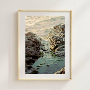 May include: A framed photograph of a rocky shoreline with a narrow channel of water leading to the ocean. The rocks are dark brown and textured, with patches of green algae. The water is a light blue, reflecting the sunlight.