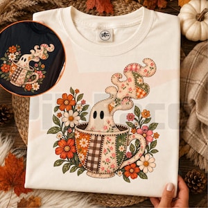 May include: A cream-colored t-shirt featuring a graphic of a ghost emerging from a patchwork-patterned teacup surrounded by orange, pink, and white flowers. The ghost and cup are decorated with floral, plaid, and gingham patterns in warm tones.
