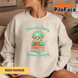 Könnte beinhalten: Cremefarbenes Sweatshirt mit einem Cartoon-Dinosaurier, der ein Buch auf einem Stapel bunter Bücher liest. Der Text "Unearth a Story" und "Summer Reading 2026" sind in Türkis gedruckt. Unten ist ein digitaler Download-Button zu sehen.