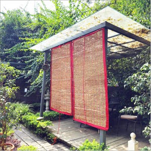 Könnte beinhalten: Zwei Bambusjalousien mit roter Einfassung hängen an einem Metallrahmen und spenden Schatten. Die Jalousien bestehen aus gewebten Bambusstreifen. Die Struktur ist von Grün umgeben, mit einer Holzterrasse.