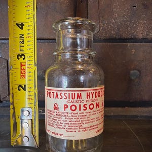 May include: A clear glass bottle with a label reading "POTASSIUM HYDROXIDE (CAUSTIC POTASH) POISON." The label includes instructions and a skull and crossbones symbol. A yellow tape measure is next to the bottle, showing a measurement of approximately 5 inches.