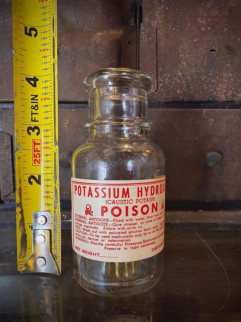 May include: A clear glass bottle with a label reading "POTASSIUM HYDROXIDE (CAUSTIC POTASH) POISON." The label includes instructions and a skull and crossbones symbol. A yellow tape measure is next to the bottle, showing a measurement of approximately 5 inches.