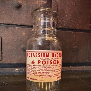 May include: A clear glass bottle with a label reading "POTASSIUM HYDROXIDE POISON." The label is red and white with black text. The bottle has a glass stopper and is likely an antique.