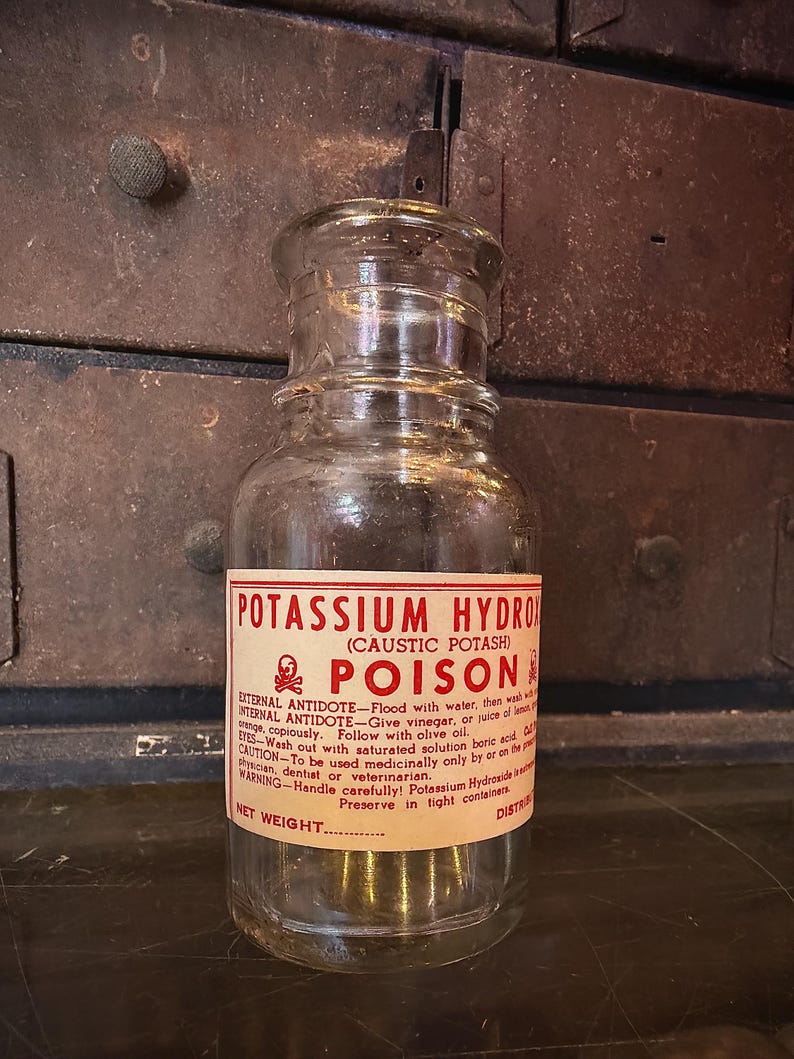 May include: A clear glass bottle with a label reading "POTASSIUM HYDROXIDE POISON." The label is red and white with black text. The bottle has a glass stopper and is likely an antique.