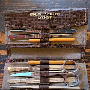 May include: A brown faux-alligator skin folding case labeled INDIANA UNIVERSITY ANATOMY containing a set of stainless steel medical dissection tools, including scalpels, scissors, and wooden-handled probes secured in individual slots.