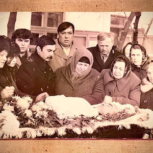 May include: Vintage black and white photograph of a funeral scene. A group of people, dressed in coats and headscarves, are gathered around a casket. The casket is covered in white fabric and surrounded by flowers. The photograph has a sepia tone.