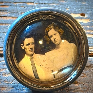 May include: A circular cabinet knob featuring a vintage black and white photograph of a person with short dark hair wearing a collared shirt and tie next to a person with wavy hair wearing a light-colored top, all encased under a clear glass dome.