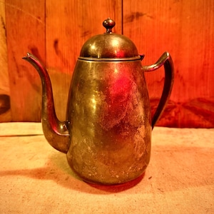 May include: A vintage brass teapot with a curved spout and handle. The teapot has a rounded body and a lid with a small knob. The brass has a tarnished appearance, suggesting age. The background is a wooden surface.