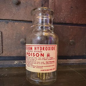 May include: A clear glass jar with a screw-on lid. The jar has a red and white label that reads "Potassium Hydroxide (Caustic Potash) POISON" with a skull and crossbones symbol. The jar is sitting on a dark surface.