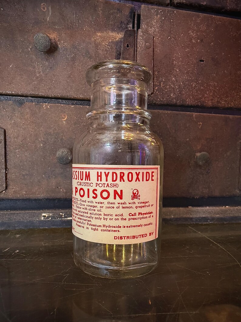 May include: A clear glass jar with a screw-on lid. The jar has a red and white label that reads "Potassium Hydroxide (Caustic Potash) POISON" with a skull and crossbones symbol. The jar is sitting on a dark surface.