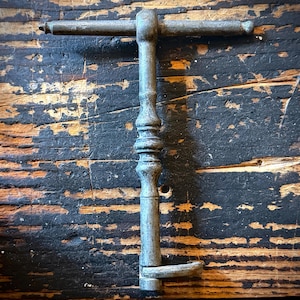 May include: A vintage metal T-handle key with a weathered gray finish. The tool features a long, slender vertical shaft with decorative turned detailing in the center and a small, L-shaped bit at the bottom end for turning mechanisms.