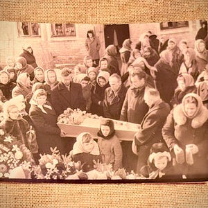 May include: Sepia-toned photograph of a funeral scene. A crowd of people, many wearing coats and headscarves, surrounds a casket adorned with flowers. The setting appears to be outdoors, near a building.