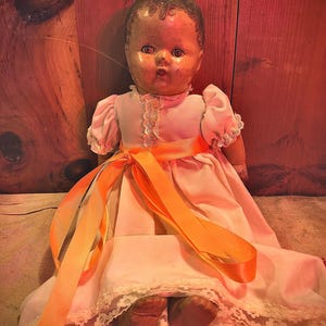 May include: Vintage doll with a painted face, wearing a pink dress with lace trim and puff sleeves. An orange ribbon is tied around the waist. The doll's feet are visible, and the background is a wooden surface.