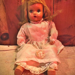 May include: Vintage doll with blonde hair, blue eyes, and a pink dress. The doll is seated, wearing a dress with a lace hem and a decorative flower. The doll's face has a painted expression.