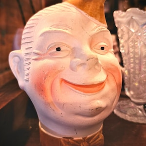 May include: A vintage ceramic head with a smiling face, painted in off-white with rosy cheeks and a tan hat. The head has black eyes and a tan collar with a bow tie. The piece has a distressed, aged appearance.