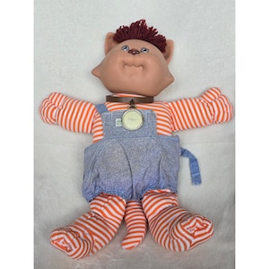 Puede incluir: Una muñeca de gato de peluche con cara bronceada, pelo de lana rojo oscuro y extremidades a rayas naranjas y blancas. La muñeca lleva un peto de mezclilla azul y un collar marrón con una etiqueta redonda de metal grabada con PIMZI.