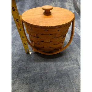May include: A round, light brown woven wooden basket with a matching lid featuring a central knob handle and a curved swing handle. A yellow measuring tape is positioned vertically against the side, showing a height of approximately 6 inches.