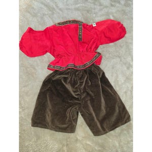 Puede incluir: Un conjunto de dos piezas que consta de una blusa roja de manga larga con puños fruncidos y un borde decorativo estampado a lo largo del cuello y la cintura, combinado con pantalones cortos de terciopelo marrón oscuro con cintura elástica.