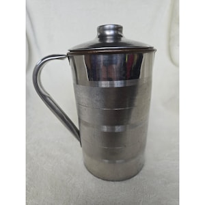 May include: A cylindrical stainless steel jug with a matching lid and a curved handle. The exterior surface features a series of horizontal brushed metal bands that wrap around the body, providing a textured appearance to the metallic finish.