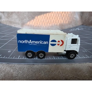 May include: A white toy semi-truck with a blue and white trailer featuring the text northAmerican VAN LINES and a blue and red circular logo. The truck has black wheels and a white cab, positioned on a gray cutting mat with visible markings.