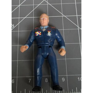 Könnte beinhalten: Eine Kunststoff-Actionfigur eines Militäroffiziers in einer dunkelblauen Uniform mit hellblauem Hemd und Krawatte. Die Figur ist auf der Brust mit aufgemalten Orden versehen, darunter ein mehrfarbiger rechteckiger Balken und eine silberne Medaille.