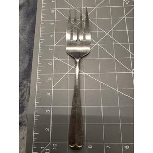 Puede incluir: Un tenedor de metal plateado con cuatro dientes colocado sobre una alfombrilla de corte gris con una cuadrícula blanca. El tenedor mide aproximadamente 19 cm de largo, desde el borde inferior de la alfombrilla hasta las puntas.