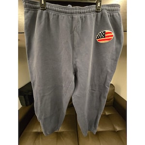 May include: Gray sweatpants with an elastic waistband and a small American flag patch on the left leg. The patch features a red and white striped design with a blue field and white stars. The pants are hanging on a hanger.