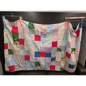 May include: A patchwork quilt constructed from numerous square fabric pieces in a variety of colors and patterns, including red, blue, green, and floral prints. The quilt is displayed hanging from two wooden hangers, showing a grid-like design.