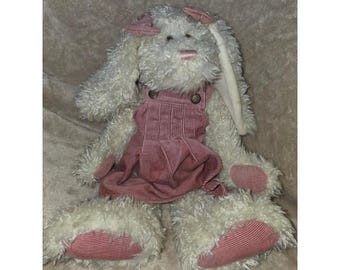 May include: A plush rabbit toy with shaggy white fur and long ears. The rabbit is wearing a dusty pink corduroy overall dress with two buttons at the chest. The rabbit has matching pink corduroy fabric on the soles of its feet and a pink nose.