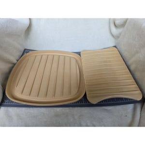 May include: A square, light tan ridged grill pan base and a rectangular, light tan ridged press lid. Both items are positioned on a dark gray cutting mat with white inch markings visible along the edges, showing the scale of the kitchenware.