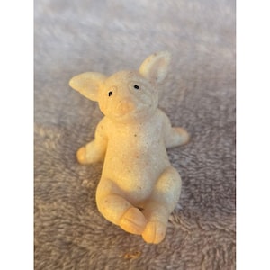 Könnte beinhalten: Eine kleine, hellfarbene Schweinefigur in sitzender Position. Das Schwein hat kleine schwarze Augen, große Ohren und seine Arme und Beine sind ausgestreckt. Die Figur besteht aus einem gesprenkelten Material und hat eine leicht raue Textur.