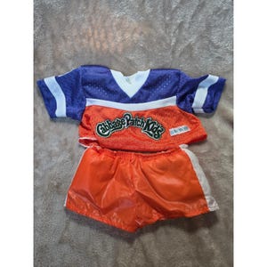 Op de afbeelding: Een tweedelige sportoutfit bestaande uit een paars en oranje mesh shirt met een witte V-hals en de tekst Cabbage Patch Kids op de voorzijde, gecombineerd met een bijpassende oranje short met witte zijstrepen en een elastische tailleband.