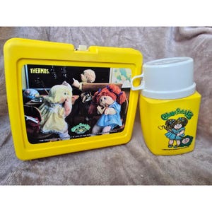 Könnte beinhalten: Eine gelbe Kunststoff-Lunchbox und eine passende gelbe Thermoskanne mit weißem Deckel. Die Lunchbox zeigt eine Grafik von Cabbage Patch Kids Puppen in einem Klassenzimmer mit dem Text THERMOS. Die Thermoskanne zeigt eine Cabbage Patch Kid Illustration.