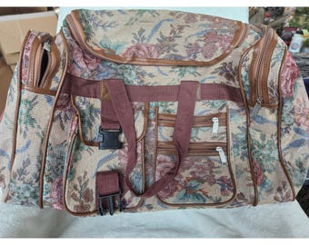 Puede incluir: Una bolsa de viaje grande con un patrón floral tejido en tonos beige, rojo, verde y azul. La bolsa incluye asas de tela marrón, múltiples bolsillos exteriores con cremallera y cierres de hebilla de plástico negro en la parte delantera.