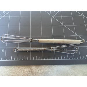 May include: Two kitchen whisks placed on a cutting mat with inch markings. The top whisk has a long, off-white handle and a wire head. The bottom whisk is smaller, constructed entirely of metal with a coiled wire handle and a wire head.