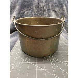 May include: A round, metallic bucket with a weathered brass-toned finish and a hinged wire handle. The bucket is positioned on a gray cutting mat with visible white grid lines and inch markings, showing a diameter of approximately 7 inches.