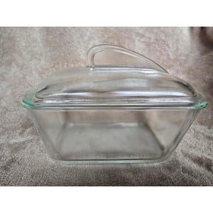 May include: A clear glass rectangular baking dish with a matching lid. The lid has a curved handle for easy lifting. The dish is made of transparent glass, allowing visibility of the contents. A classic kitchenware item.