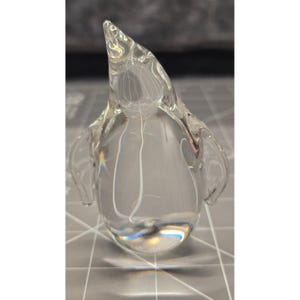 May include: A clear glass figurine shaped like a penguin. The object is made of transparent, smooth glass with a rounded body, a pointed head, and two small, wing-like protrusions on the sides. The surface reflects light, creating highlights.