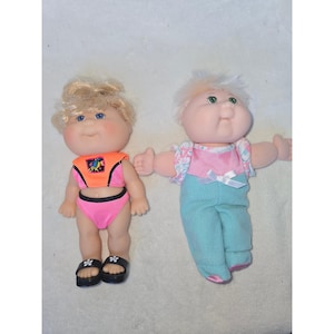 May include: Two dolls with round faces and yarn hair. The left doll has blonde hair, blue eyes, and wears a bright orange and pink swimsuit with black sandals. The right doll has light hair, green eyes, and wears a pink and teal jumpsuit.