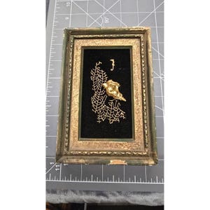 Puede incluir: Una pieza de arte mural rectangular que presenta una concha, un abanico de mar y una pequeña figura de caballito de mar pintados en dorado sobre un fondo de terciopelo negro, dentro de un marco de madera desgastado en tonos oro y verde.