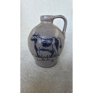 Puede incluir: Una jarra de gres gris claro moteado con una ilustración pintada a mano en azul oscuro de una vaca sobre la hierba. La jarra tiene un cuerpo redondeado, un cuello estrecho y un asa curva unida al cuello y al hombro de la pieza.