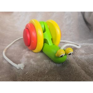 以下が含まれることがあります： カラフルな木製の引っ張りおもちゃのカタツムリ。 カタツムリの体は緑色で、黄色と赤の円形の殻のセグメントがあります。 黄色の目と白い引き紐が付いています。 クラシックな子供のおもちゃです。