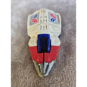 May include: A vintage toy in the shape of a claw, featuring a white body with red and blue accents. The claw has silver-colored tips and is designed with a retro aesthetic. The toy has a rectangular blue center and two red sections.