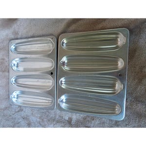 May include: Two rectangular silver-colored metal baking pans, each featuring four elongated, recessed molds shaped like corn cobs. The pans have a smooth, metallic finish with distinct ridges within each mold to create the texture of corn kernels.