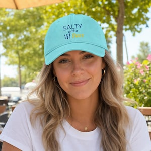 Puede incluir: Una gorra de béisbol de color turquesa claro con el texto SALTY with a 'lil Sass bordado en letras azul oscuro y amarillo en la parte delantera. La gorra cuenta con una corona estructurada, una visera curva y un botón en la parte superior.