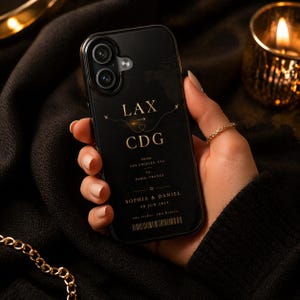 Könnte beinhalten: Schwarze Handyhülle mit goldfarbenem Text, der "LAX CDG" anzeigt, sowie Flugdetails von Los Angeles nach Paris. Die Hülle wird in einer Hand gehalten, ein goldener Ring ist sichtbar. Klare Umrandung.