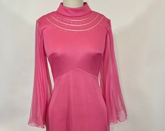 May include: A vintage pink maxi dress with a high neckline and long, sheer sleeves. The dress features a fitted bodice and a flowing skirt. The neckline is embellished with a delicate beaded design. The dress is displayed on a mannequin.