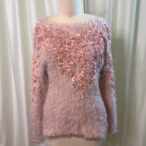 May include: A light pink, long-sleeved sweater with a fluffy texture. The sweater features a boat neck and is adorned with delicate, petal-like embellishments across the chest and sleeves. The sweater is displayed on a mannequin.