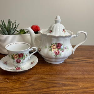 May include: A white porcelain tea set comprising a teapot, teacup, and saucer. The set is adorned with a floral rose pattern in red, yellow, and pink. The teapot has a lid and a curved handle. The teacup and saucer also feature the rose design.