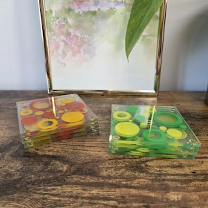 Peut inclure: Deux piles de dessous de verre carrés en matériau transparent. Une pile présente un motif de cercles rouges, orange et jaunes de tailles variées. L'autre pile présente un motif de cercles vert clair, vert foncé et citron vert de tailles variées.
